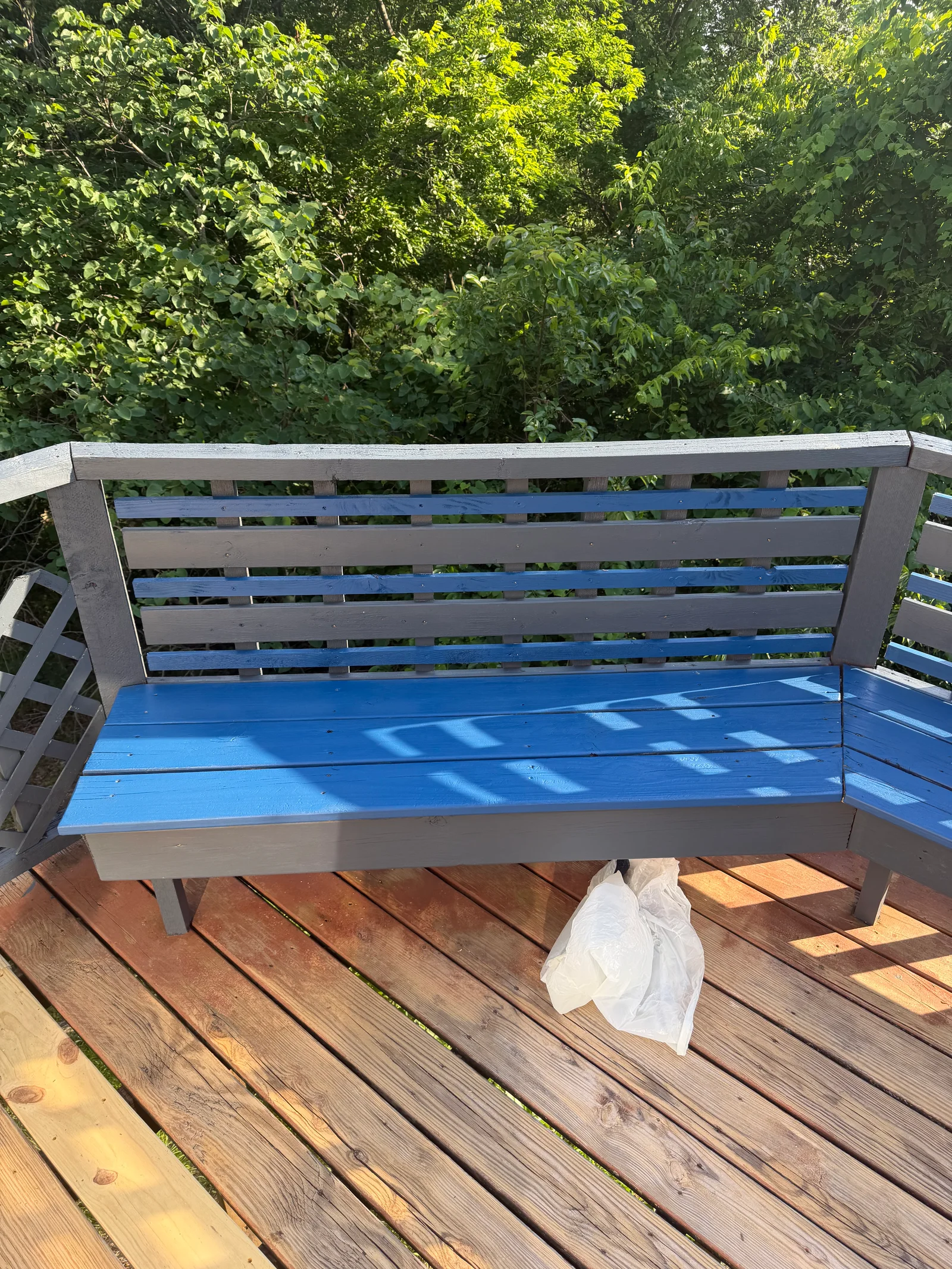 Blue painted wooden bench with gray frame on brown deck, surrounded by green foliage