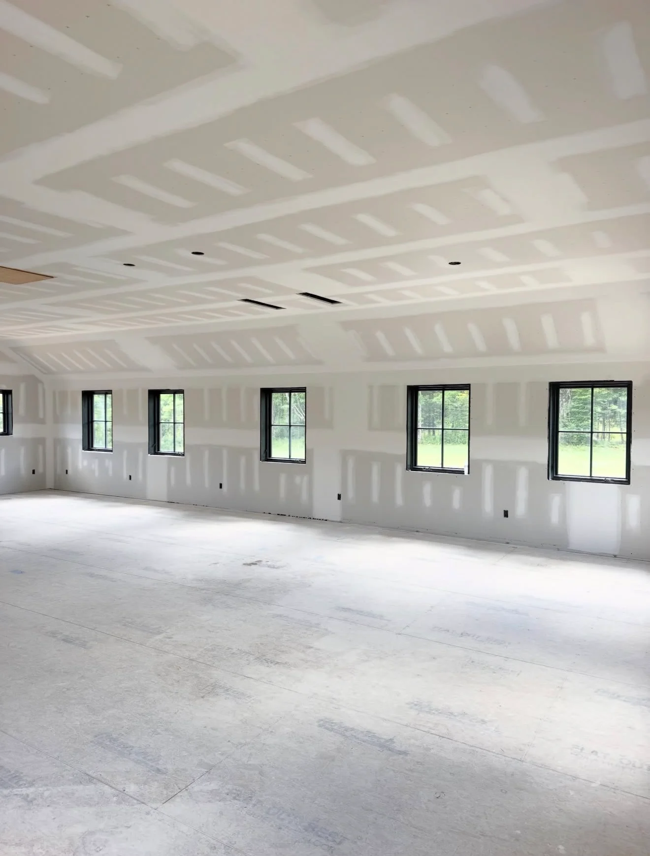 Drywall finishing with taped seams, black windows, concrete floor, and exposed ceiling joists